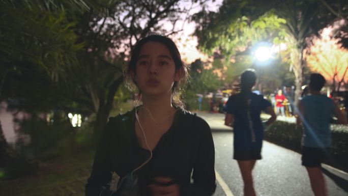迷人的亚洲少女一边在公园跑步一边听音乐。年轻女子在夜间慢跑锻炼。