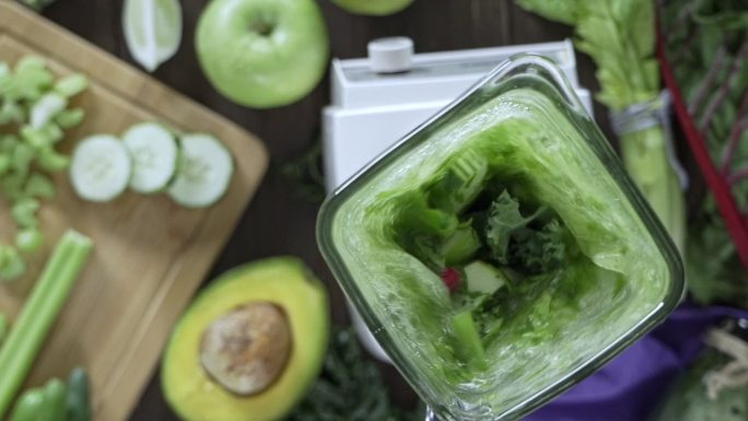 饮食概念：慢动作的直接下方镜头的搅拌机混合蔬菜和水果排毒冰沙。餐桌上的砧板和搅拌机混合