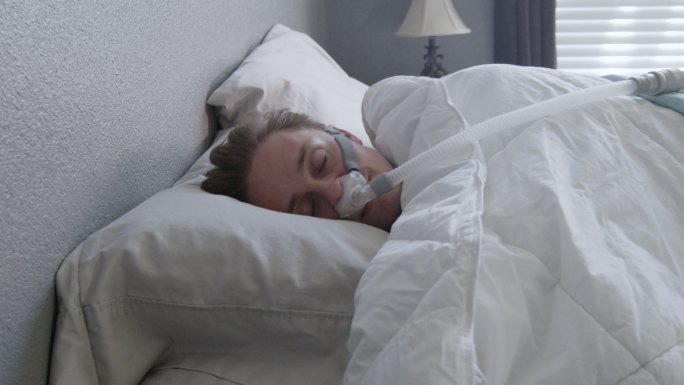 一名患有睡眠呼吸暂停症的年轻男子侧躺在床上，戴着CPAP面罩，特写镜头