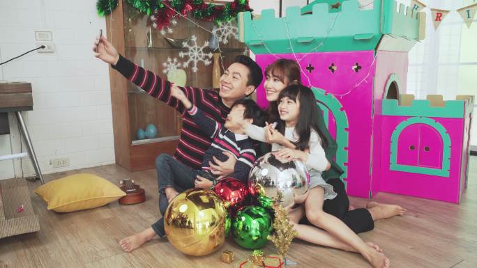 一个亚洲家庭坐在地板上和纸做的家庭玩具一起准备圣诞节。12月的假期，父亲用智能手机拍摄集体自拍照片，