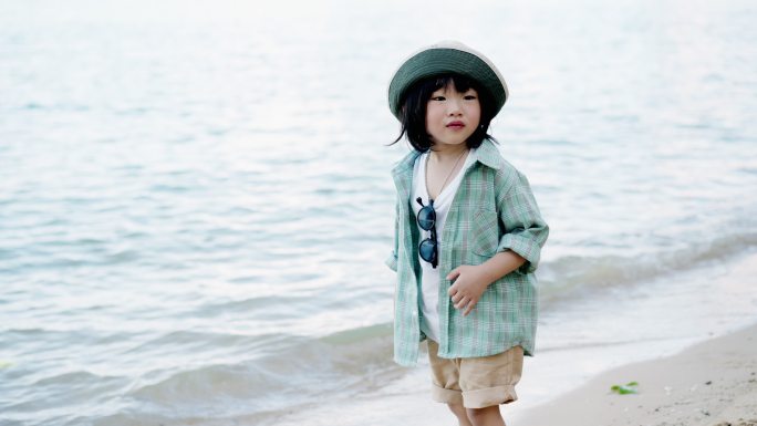 穿着绿色格子呢衬衫的快乐亚洲男孩在海滩上散步，寻找贝壳，看着海水