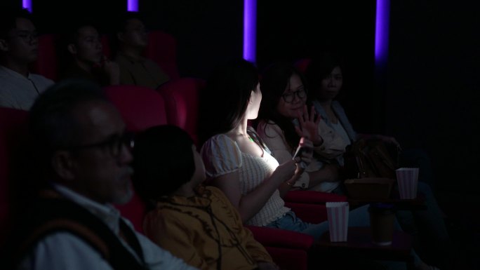 中国亚裔年轻女子在电影院放映电影时使用智能手机。打扰她周围的其他观众