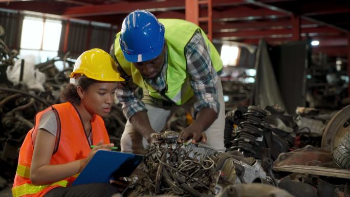 戴着安全帽的多种族工人使用剪贴板检查车辆废料场的备件。