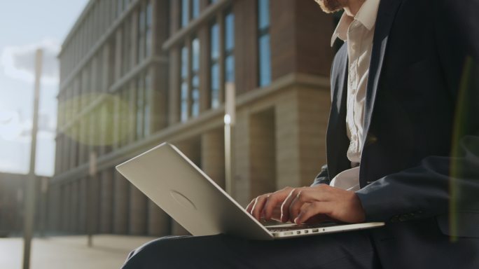 一个穿着深蓝色西装和白色衬衫的年轻商人在美丽的阳光下坐在现代建筑的旁边，在笔记本电脑上打字，脸上挂着