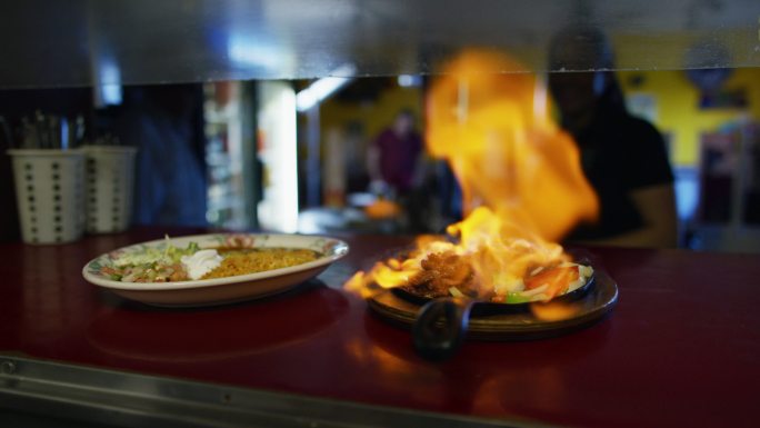 一名厨师在一家墨西哥餐厅的服务窗口点燃一盘法吉塔（Flambe），一名女服务员走了过来