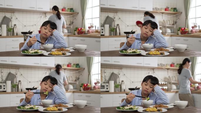 一个心烦意乱的亚洲学龄前男孩在餐桌上拿起碗里的蔬菜，脸上带着仇恨的表情，而他的母亲正在厨房里做饭