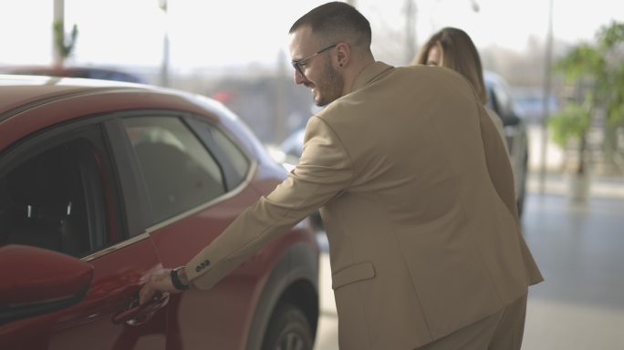 女销售员和男客户走近汽车并进入，而女销售员则在汽车经销商处向其展示汽车内饰