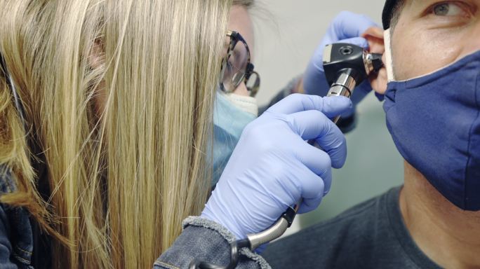 在一家诊所的检查室里，一名戴着外科口罩的女医生助理（PA）用耳镜检查一名40多岁戴面具的白人男子的耳