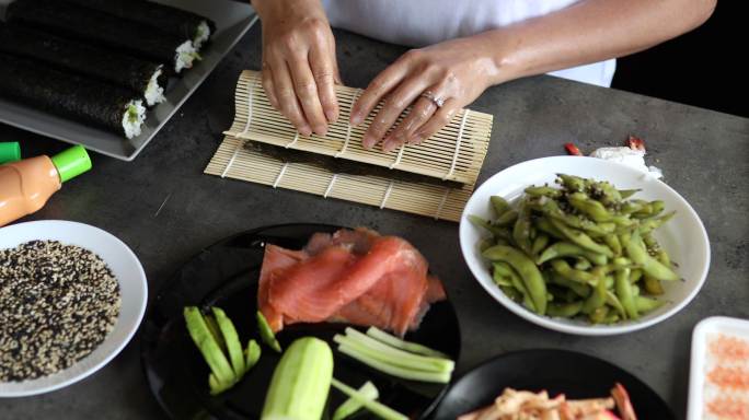 熟练的女士用竹垫卷起传统寿司，寿司由黄瓜、鳄梨和熏鲑鱼制成