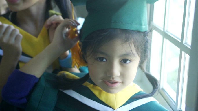 身穿毕业帽和毕业礼服的亚洲小学生站在学校里微笑。