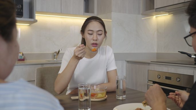 一名穿着睡衣、喉咙痛的亚洲女子正在家中厨房的桌子上吃鸡汤以缓解喉咙痛，并与朋友交谈