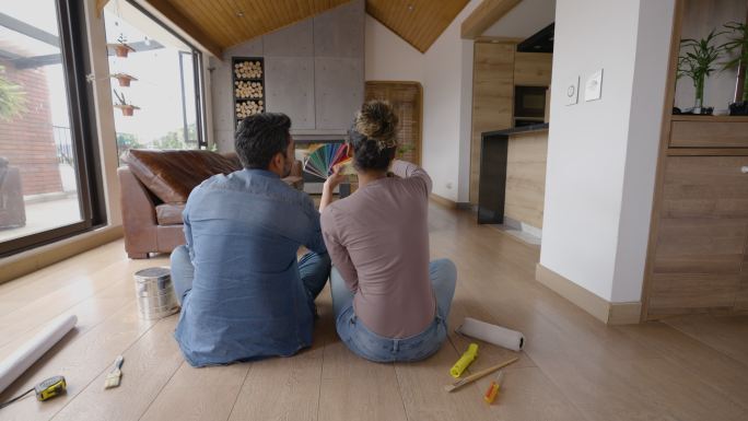 不同的夫妇坐在地板上，为新家的墙壁选择不同的颜色-后视图照片