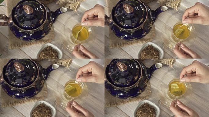 白色乡村桌子上，一位女士双手将茶包搅拌到热茶甘菊输液杯中的高角度特写镜头