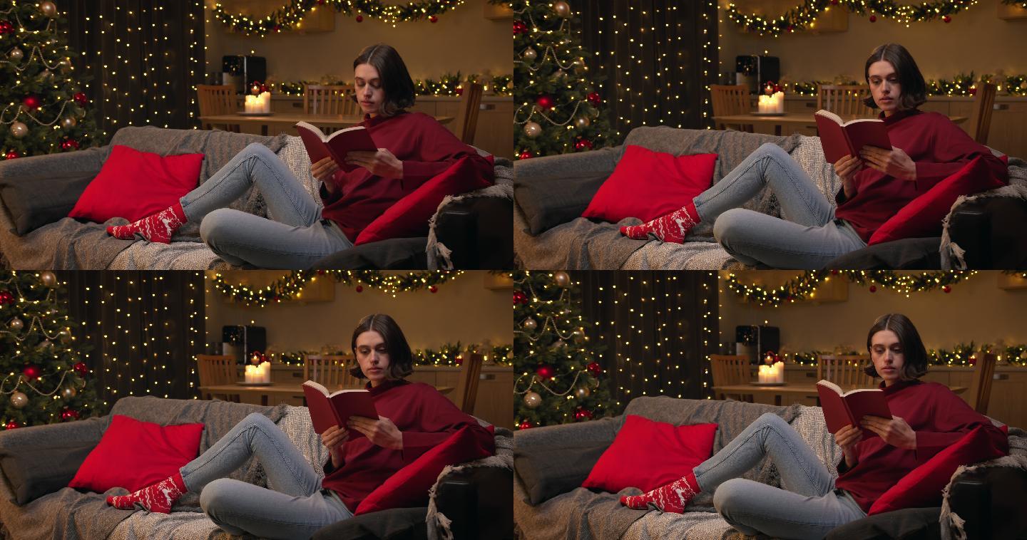 一位穿着红色毛衣和牛仔裤的年轻迷人的女士坐在一间舒适的圣诞装饰房间的沙发上，读着一本有趣的书