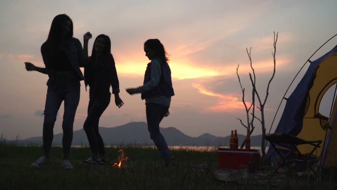 低角度视图：朋友在夏天围着营火跳舞，日落时在湖边露营