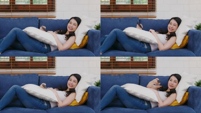 年轻的亚洲女性一边看电视一边拿着遥控器躺在家客厅背景的沙发上，人们享受着生活方式
