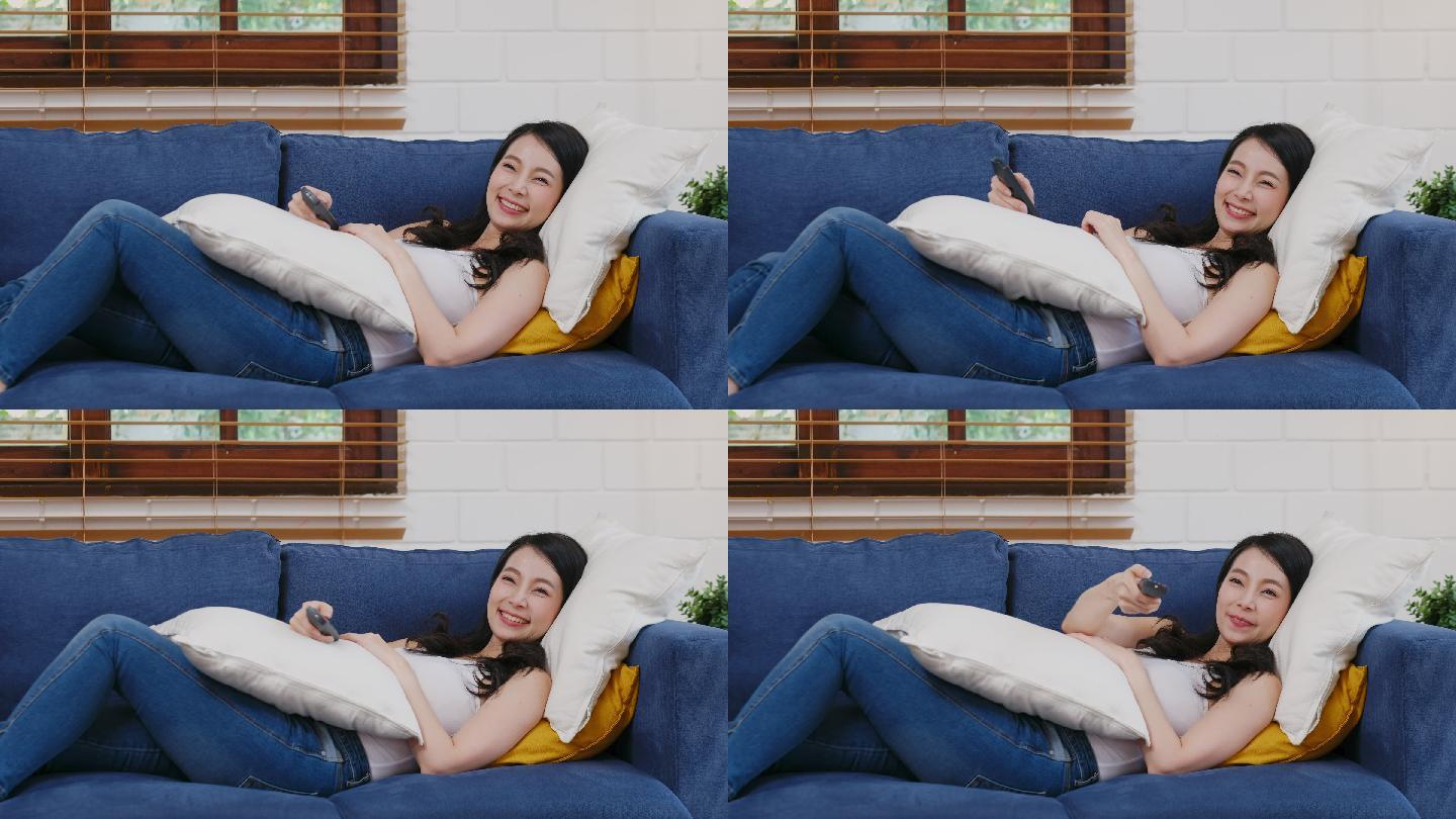 年轻的亚洲女性一边看电视一边拿着遥控器躺在家客厅背景的沙发上，人们享受着生活方式