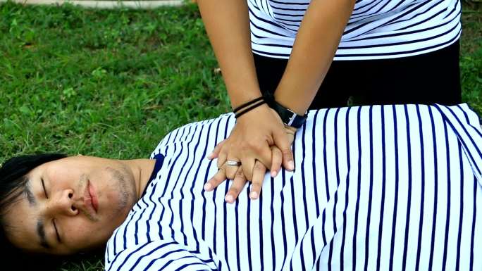救生员CPR培训穿t恤条纹的男子躺下，穿白色长袖衬衫的女子在草地上培训CPR背景，复苏过程中的概念（