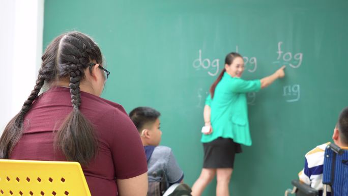 青少年唐氏综合症女孩在轮椅上与残疾学童小组练习阅读、发音英语单词，亚洲美女老师站着给予鼓励，感受幸福