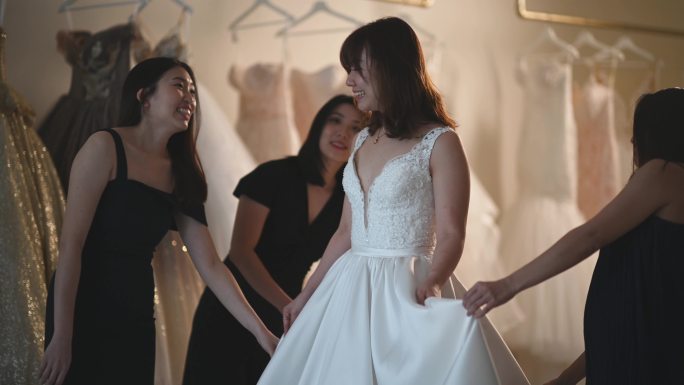 亚洲华人新娘在伴娘的簇拥下试穿婚纱，在婚纱店欣赏婚纱的美丽