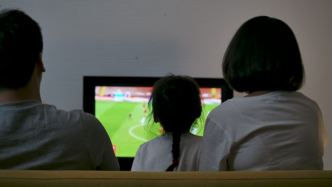 一个亚洲家庭坐在沙发上看电视足球比赛，一起欢呼，为胜利而高兴。