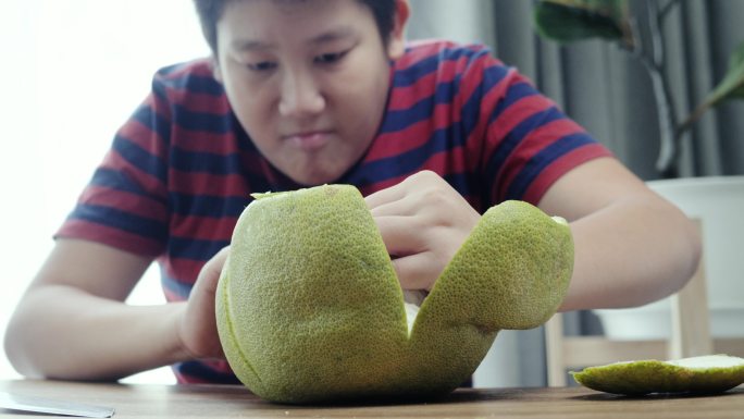 快乐的亚洲男孩坐在家窗户附近的地板上剥柚子，生活方式理念。