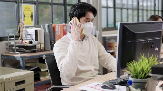 新常态的环境在现代办公室工作带有2019冠状病毒疾病，亚洲商人通过屏幕分隔与客户和朋友打电话做生意，