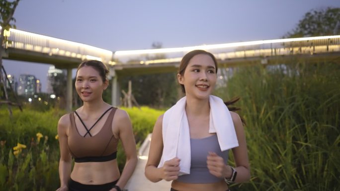 正面图：两位亚洲美丽的中年女性朋友穿着运动装，肌肉发达，在黄昏时分一起积极慢跑，使用智能手表监控，在