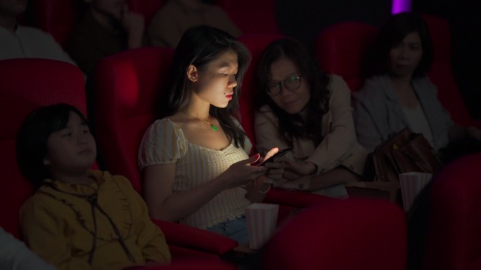 一名亚裔中国女性在电影放映期间，在黑暗中阅读手机短信，忽视并干扰周围的其他观众