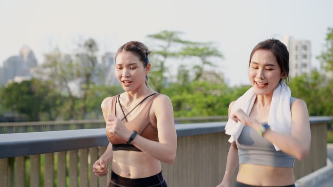 两位亚洲美丽的中年女性朋友穿着运动装，肌肉发达，早上在市中心附近公园花园的人行桥上，一起积极慢跑，用