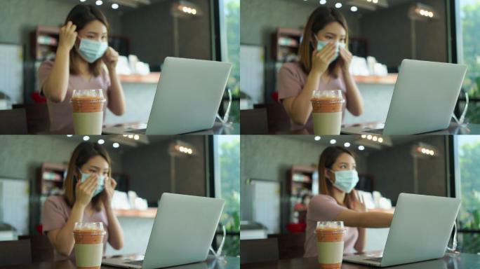 一家小咖啡馆再次开业后，亚洲自由职业女性戴着防毒面具坐在那里，用笔记本电脑工作。