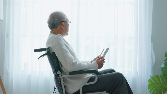 亚洲老年男性独自坐在轮椅上看家庭照片。年迈、成熟的祖父感到孤独和悲伤，退休后留在养老院思念亲人和妻子