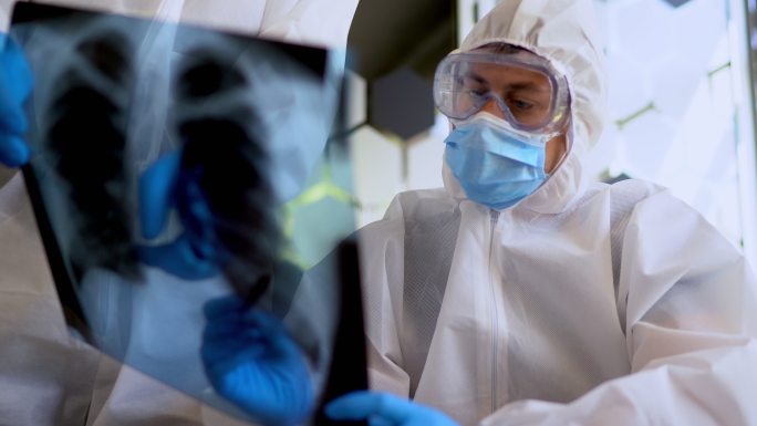 穿着流行病学防护服的医生正在检查患者肺部的X光片