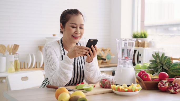 亚洲资深女性在准备自制不同口味的水果冰沙和酸奶时，使用手机感到很开心。健康的天然益生菌风味饮料，学习