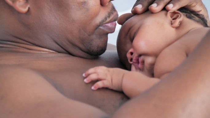 特写镜头中，幸福的非洲父亲在3个月大的新生儿睡在他的胸脯上时拥抱亲吻。家庭时刻概念。