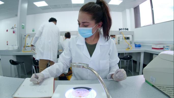 在生物实验课上，一名女生在显微镜下观察样本，用笔记和智能手机上的视频记录一切