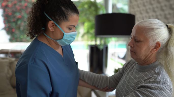 家庭护理人员帮助一位老年女性在家站立-医生戴着防护面罩
