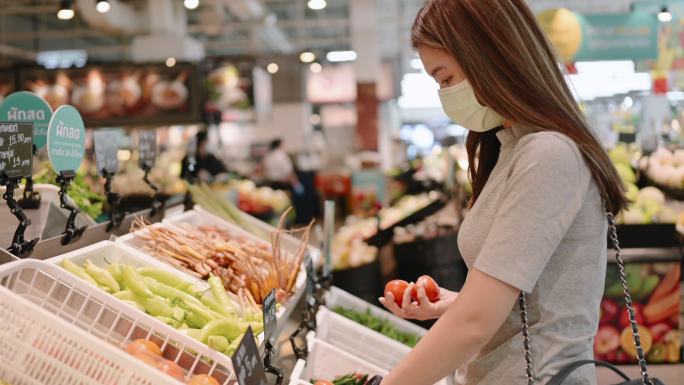 社交距离概念亚洲女性戴着防护面罩，晚上在超市选择西红柿、蔬菜和水果
