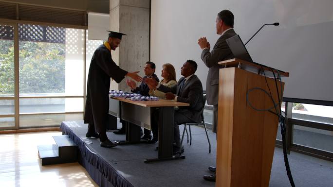 英俊的男研究生在领取大学文凭时与董事会握手