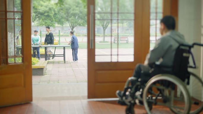 一名坐在轮椅上的少年看着窗外玩耍的伙伴