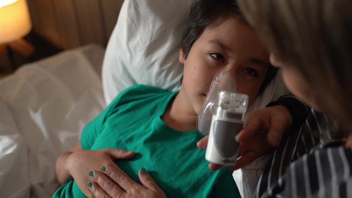 母亲帮助儿子在家床上进行吸入治疗时使用雾化器