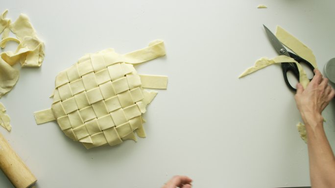 一个女人的手在头顶上将一条条糕点面团编织在一起，形成一个格子状的馅饼表层，然后用剪刀修剪边缘
