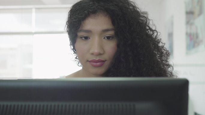 一位留着非洲头发的拉丁女性坐在办公室的电脑前，为客户工作