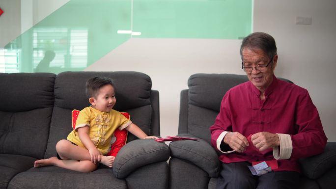 中国农历新年期间，在客厅打包红包的亚裔中国人微笑可爱的孙子看着爷爷