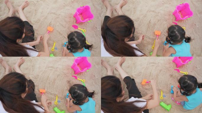 亚洲可爱的蹒跚学步的小女孩坐在沙滩上，和妈妈一起玩沙滩城堡。