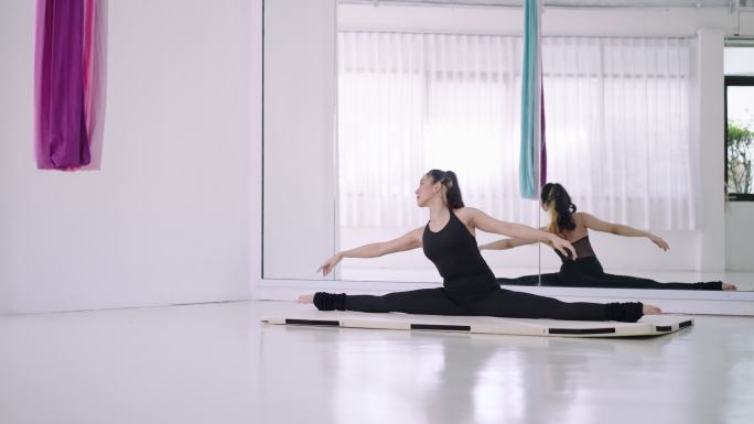 一位年轻的亚洲女性在有镜子的白色房间里，在瑜伽垫上以劈开的姿势伸展双腿。练习瑜伽锻炼身体的灵活性和伸