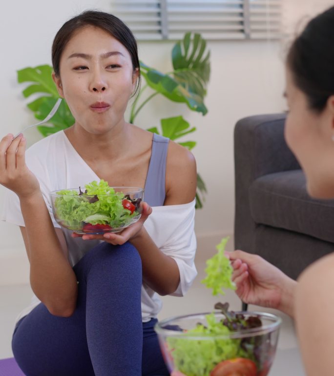 两位穿着运动装的美丽亚洲年轻女性手里拿着一杯新鲜沙拉和一把叉子。节食的概念素食女性吃健康美味的食物。