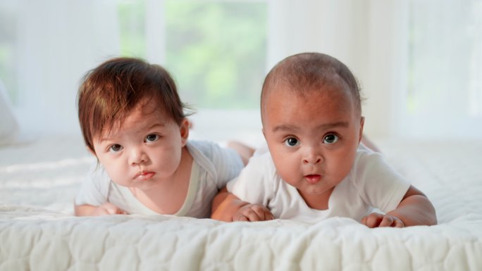 一个无辜的两个穿着白色婴儿服的黑白相间的新生儿躺在床上，试图爬行并看着摄像机。