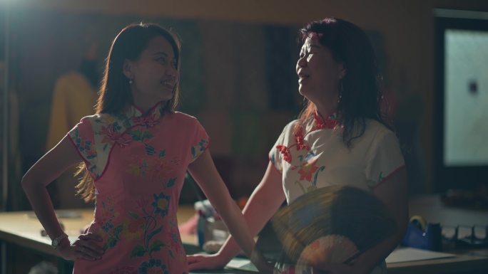 亚洲华人女裁缝小企业主和她的母亲站在工作场所，穿着中国传统服装旗袍，手持折扇，双手放在下巴上，自信地