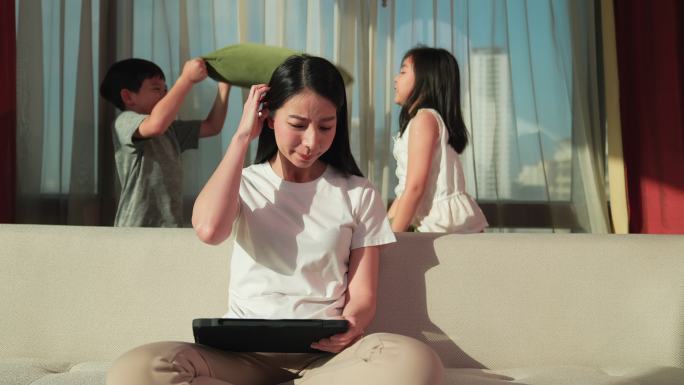 亚洲母亲在家里制作平板电脑。被儿子和女儿打扰的她正在享受枕头大战。使用那些不得不在家工作并有孩子的人